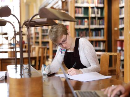 A student in the library. There are bookshelves behind them and they are studying at a desk.