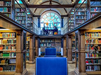 A blue chair in the middle of a library. There are bookshelves on either side and an arched and decorated cieling above it. 