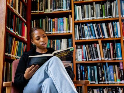 A student sitting in a library reading a book. There are bookcases all around them. 
