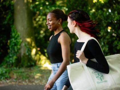 Two people walking. They are smiling and one is holding a large tote bag. There are trees behind them