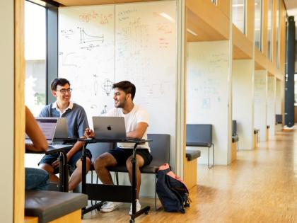 Two students sitting on Apple macs. They are in a long corridor filled with natural light. 
