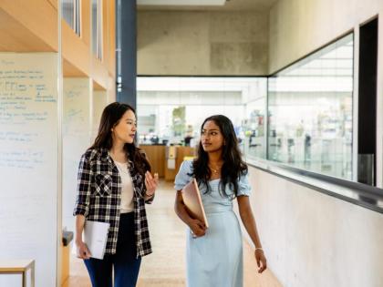 Two students walking down a corridor. One is carrying a book and the other is holding a laptop