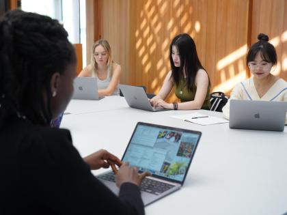A group of three students are sitting on their laptops at a shared table. In the foreground is another student on their laptop typing