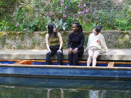 Three students sitting on a riverbank. They have their feet in a blue boat that is floating on the water. 