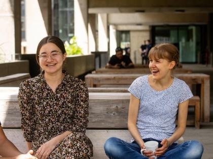 two students sitting on a benching. They are laughing and one is drinking coffee. 