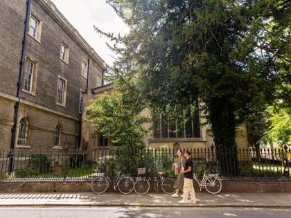 A student walking along a pavement. Behind them is a fence with bikes leaning on it, a tree and a building. 