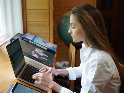 A student with their hands on a laptop keyboard. They are working on a wooden desk facing a window.