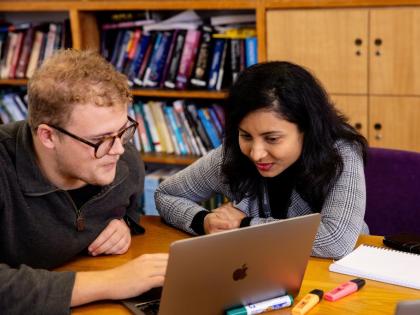 Two students on an Apple laptop. There are books on the shelf behind them. 
