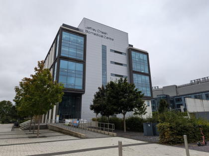 The Jeffrey Cheah Biomedical Centre that is grey with large glass windows. There are trees in the foreground.