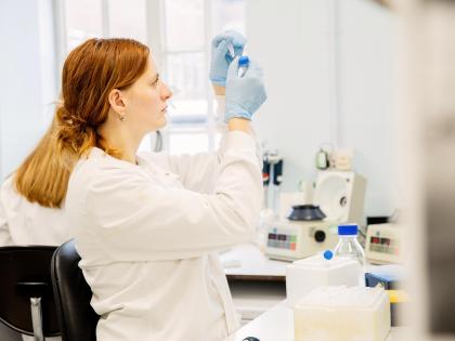 A person in a lab. They are wearing a lab coat and are holding up a sample to look at. Behind them is a variety of lab equipment. 