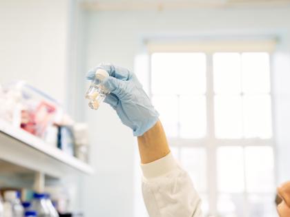 A gloved hand is holding a test tube up in the air. Behind them are some shelves and a window. 